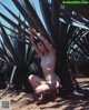 A naked woman kneeling in front of a palm tree.