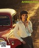 A woman leaning against a red car on a dirt road.