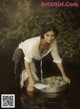 A woman in a white shirt and brown skirt is washing clothes in a metal tub.